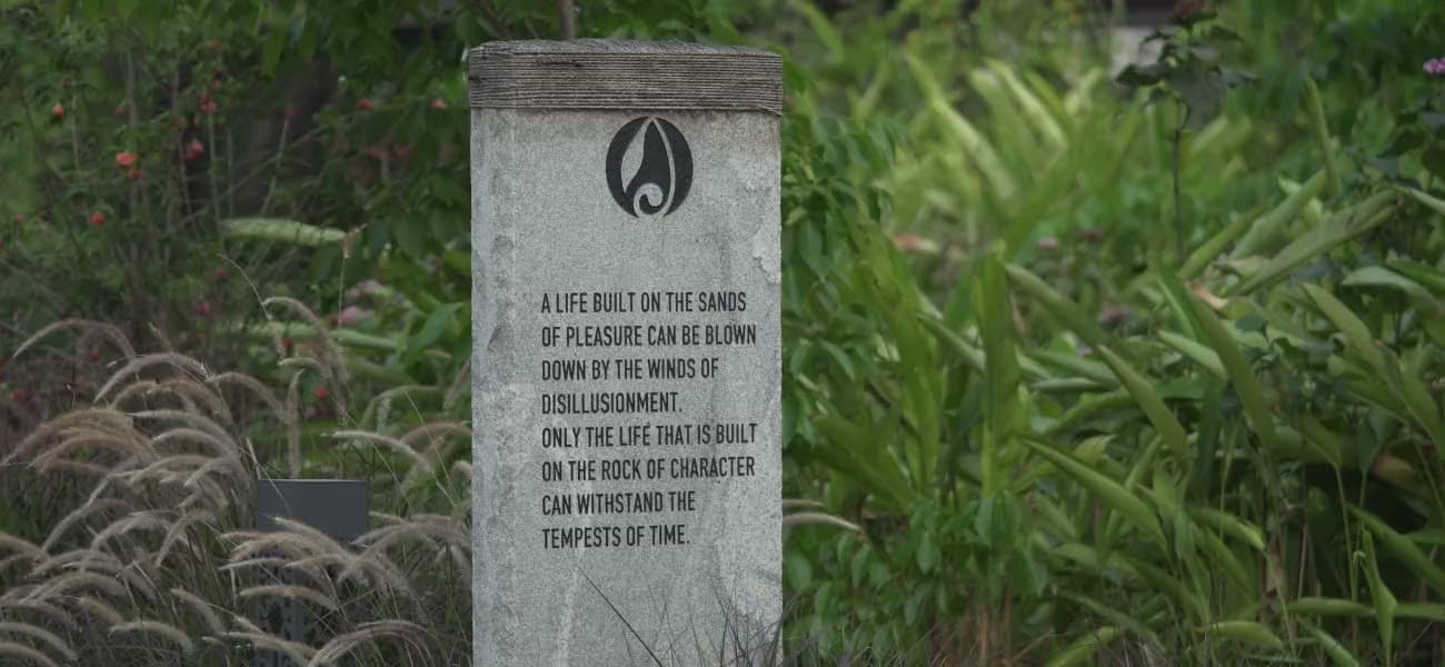 A stone marker set among tall grasses, inscribed with the Alekhya mark and the words: a life built on the sands of pleasure can be blown down by the winds of disillusionment; only the life that is built on the rock of character can withstand the tempests of time